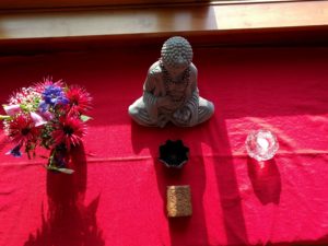 A small stone Buddha flanked by a small summer bouquet of flowers and a tealight candle inside a rough glass container.